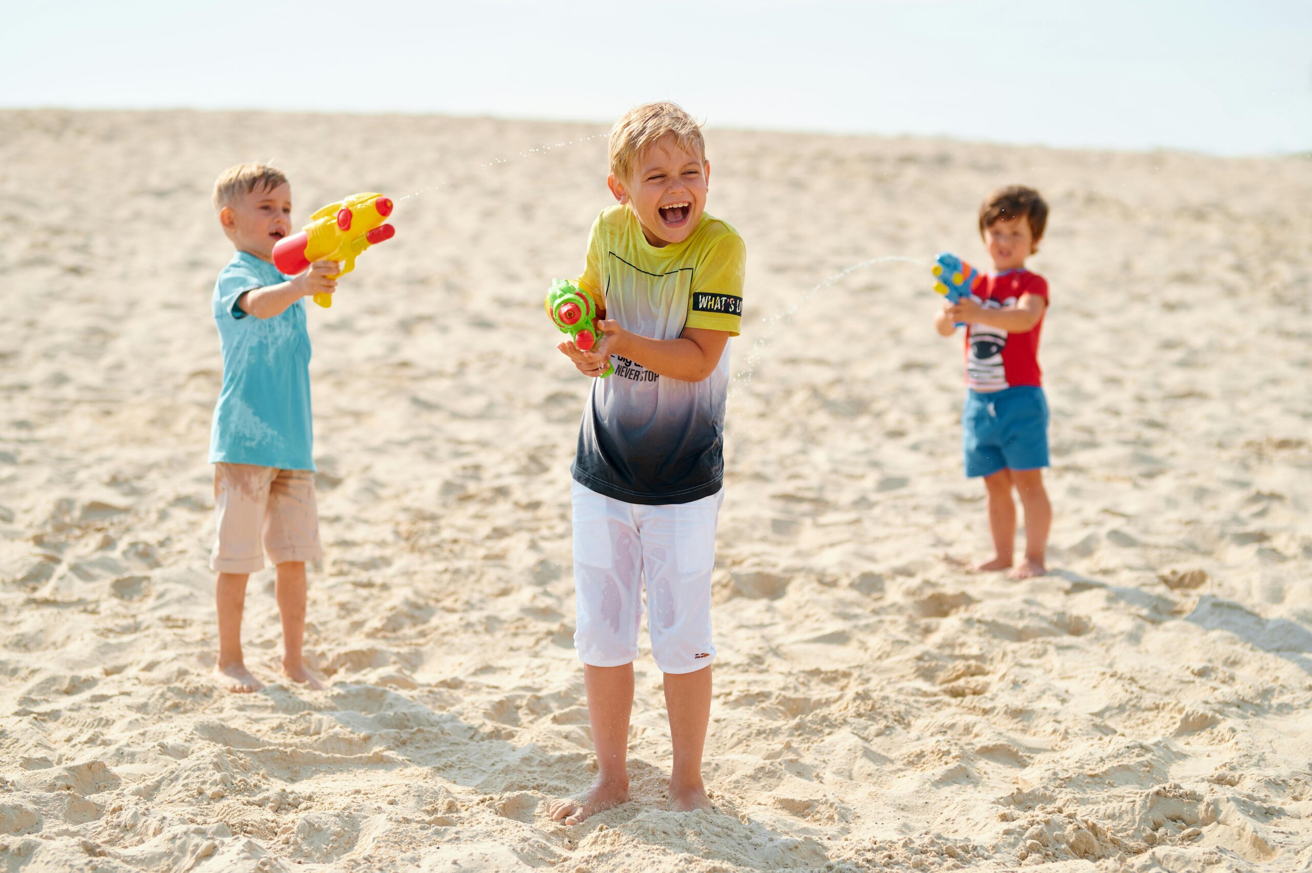 Three children joyfully playing with water guns on a sandy beach under the sun.